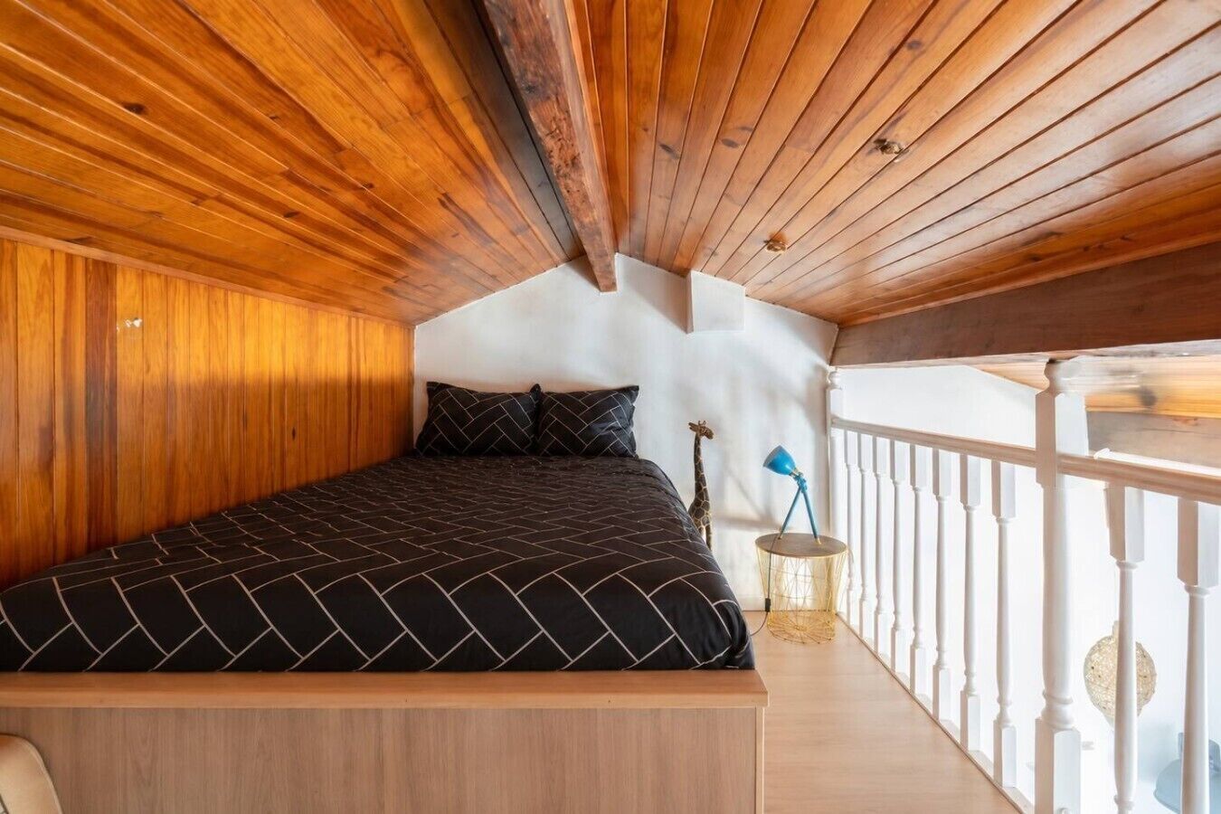 A bedroom with a wooden ceiling, a bed with a black and white patterned bedspread, a giraffe decoration, and a white railing.