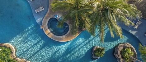 An aerial view of a resort with a central swimming pool, lounge chairs, and palm trees.
