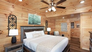 Un dormitorio tipo cabaña de madera con cama, mesitas de noche, ventilador de techo y decoración en las paredes.