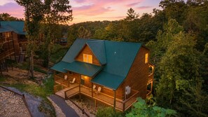 A cabin with a porch, multiple windows, and a green roof, surrounded by trees. - Mountain High | Spacious+Hot Tub+Game Room+More! (Pigeon Forge)
