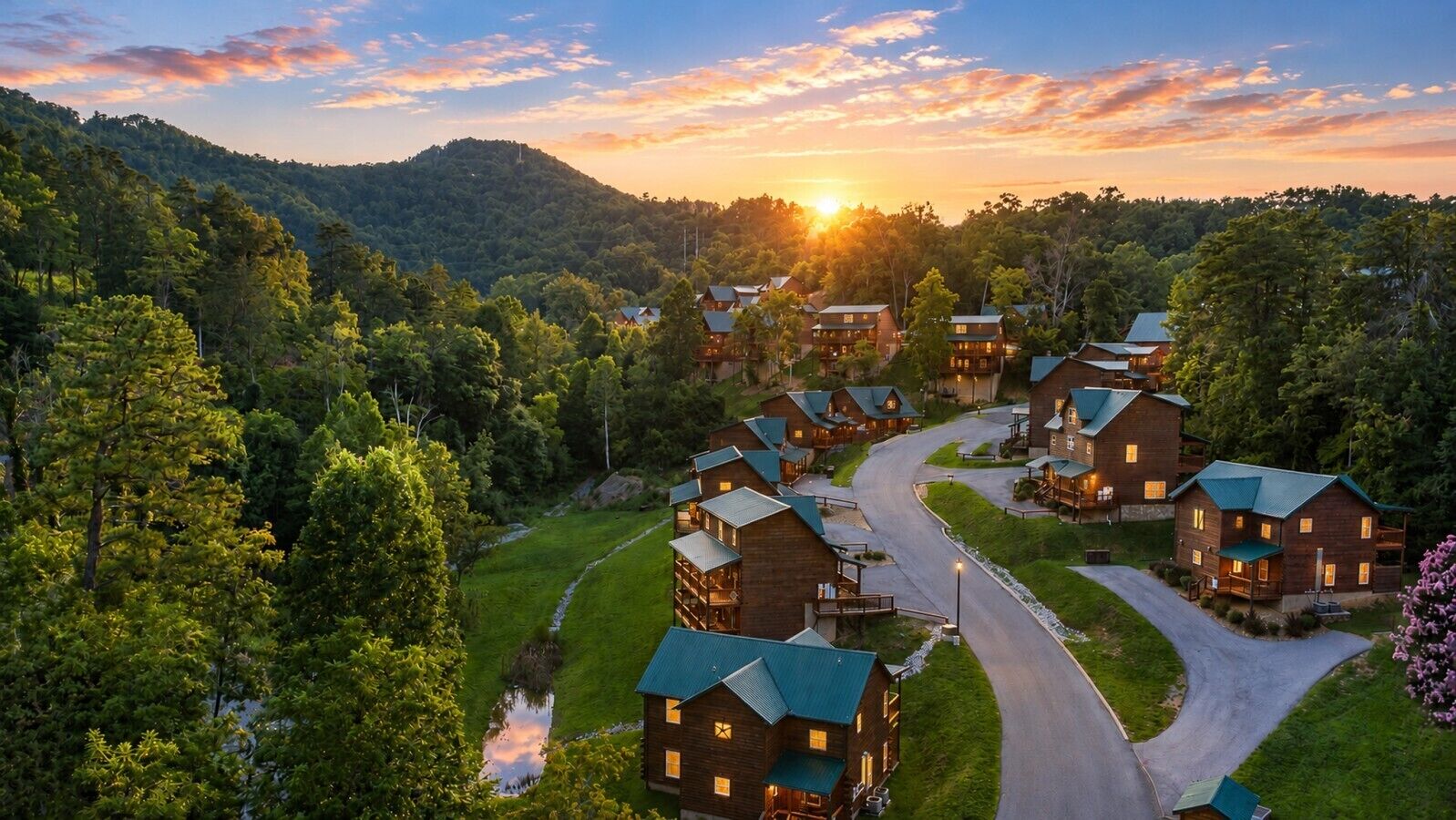 A cluster of log cabins with a winding road connecting them, surrounded by dense forest and hills.