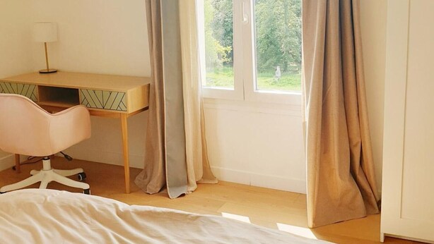 A bedroom with a bed, a desk, and a chair. There is a window with curtains.