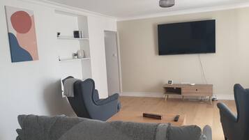 Una sala de estar con un televisor de pantalla plana montado en la pared, una mesa de centro de madera y dos sillones grises.