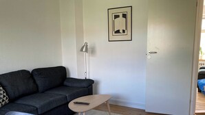Ein Wohnzimmer mit einem dunkelgrauen Sofa, einem kleinen runden Tisch mit Fernbedienung, einer Stehlampe und einer weißen Tür mit Schlüsselloch.