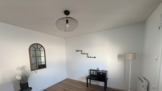 Una stanza con pavimento in legno, un tavolo nero con una sedia, uno specchio a parete, un lampadario e un termosifone.