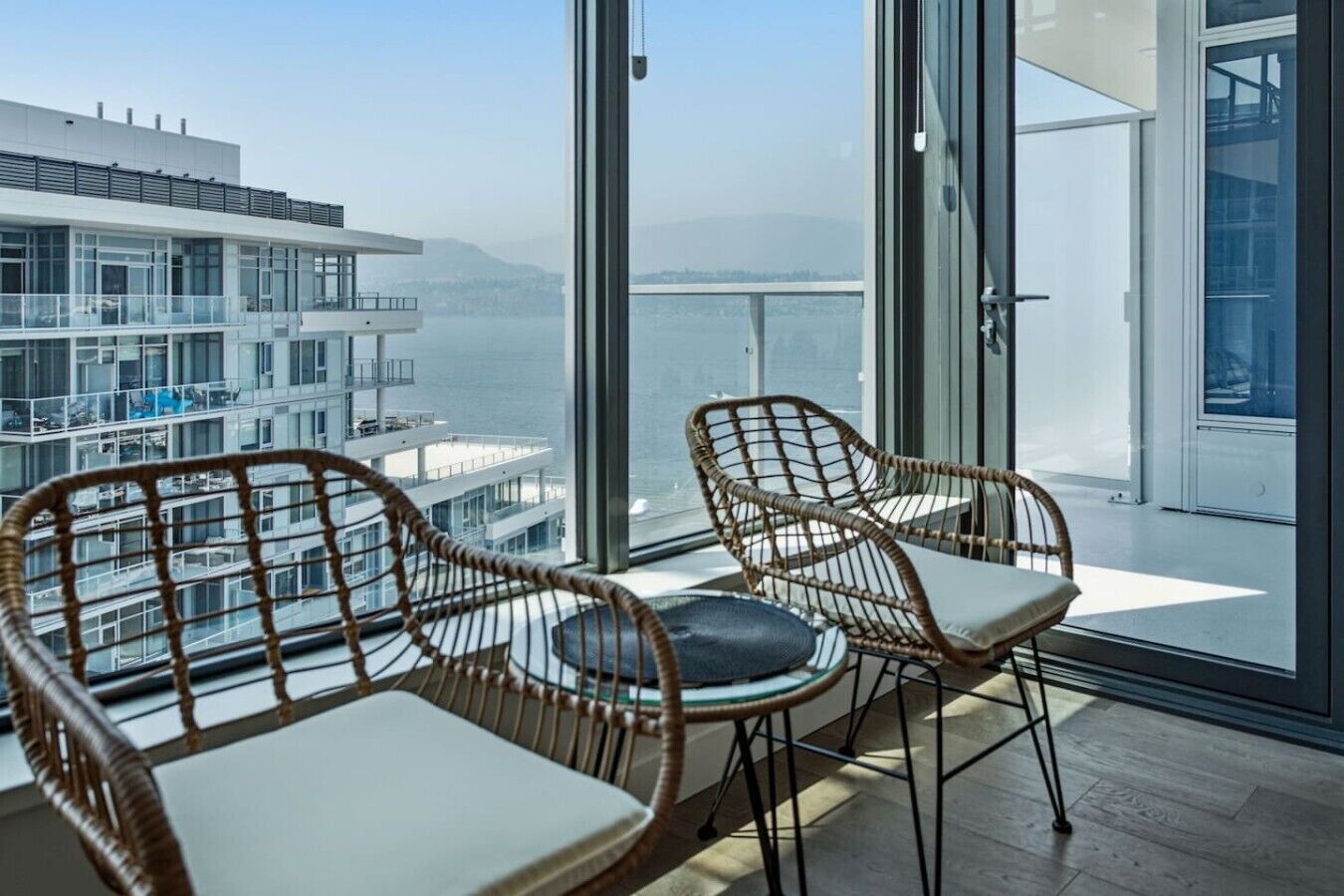 Un balcón con sillones de mimbre y una mesa, con vistas a un edificio moderno y una masa de agua.