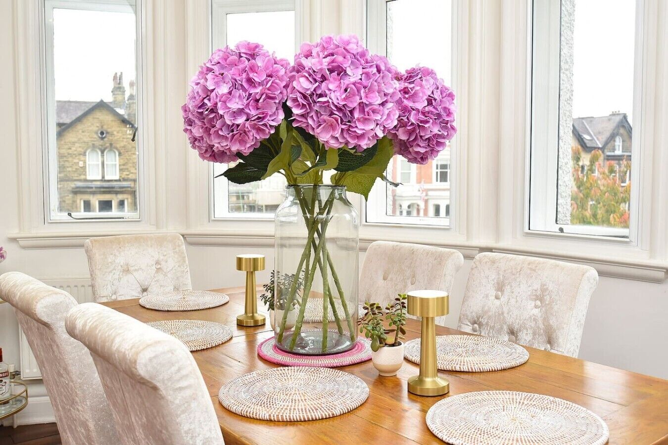 A dining table with a vase of flowers, surrounded by chairs, in a room with large windows.