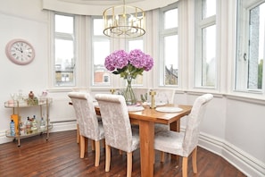 A dining area with a wooden table, six chairs, a vase of flowers, a clock, and a window with a view of buildings. - Beautiful & Luxury Apartment Overlooking The Stray (North Yorkshire)