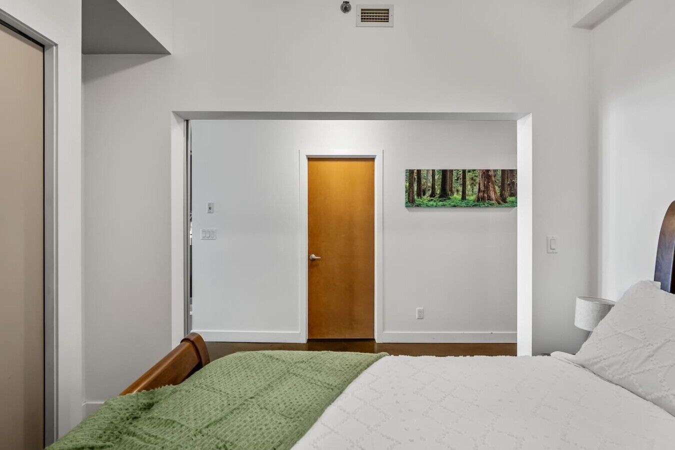 Un dormitorio con una cama, una manta verde, un cuadro de un bosque y una puerta que da a otra habitación.
