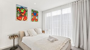 Un dormitori amb un llit, cortines blanques i dos quadres de colors a la paret.