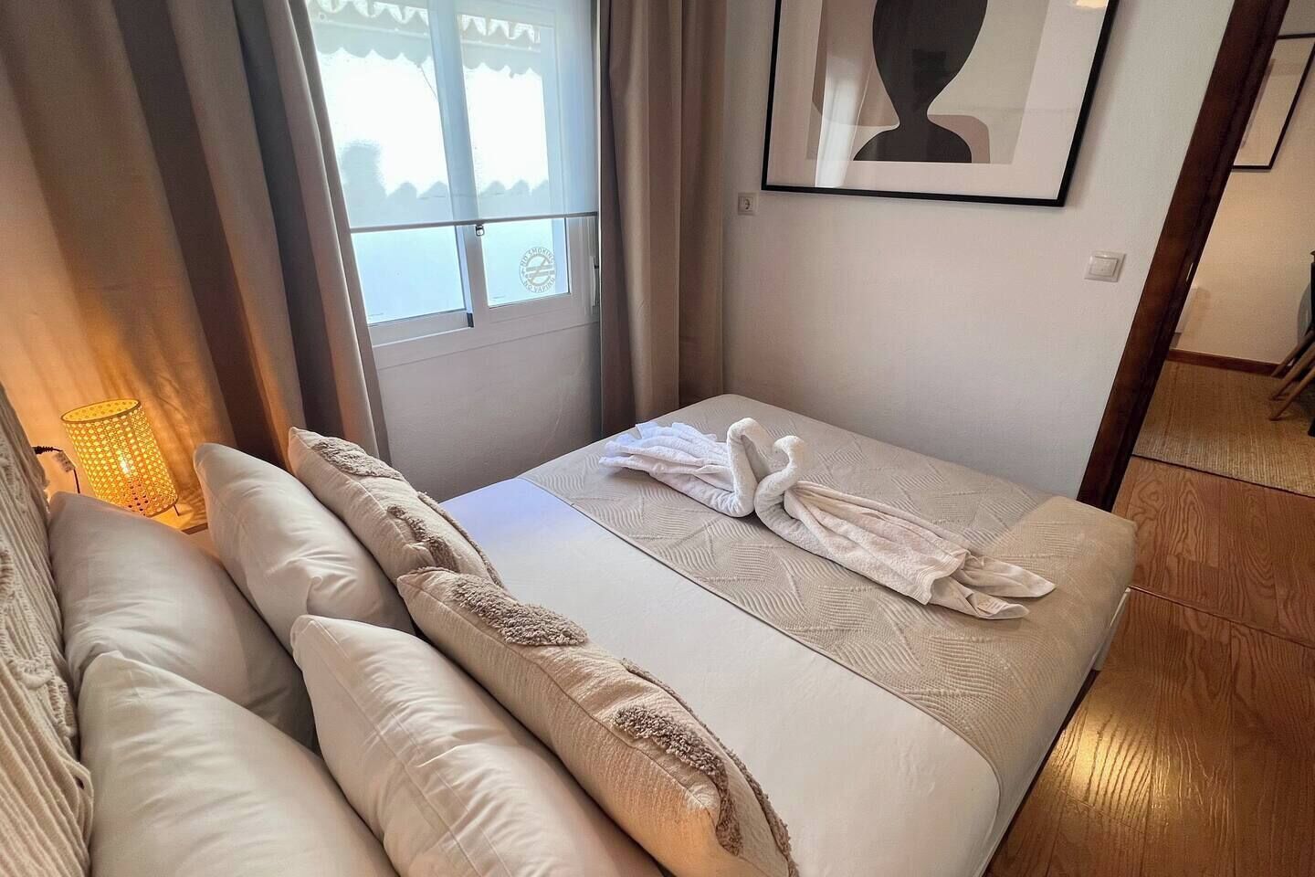 A hotel room with a bed, pillows, a lamp, a window, and a framed artwork on the wall.