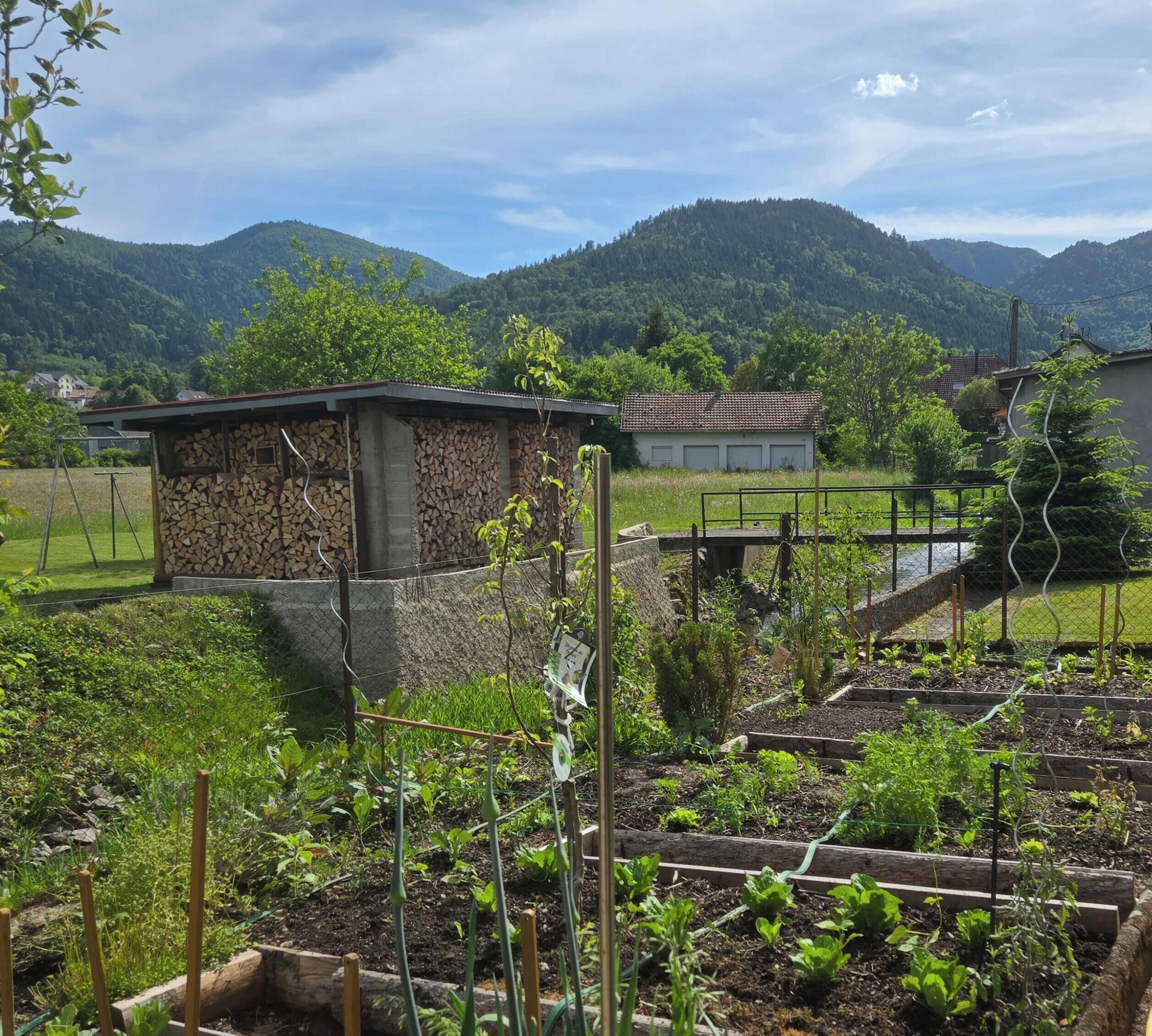 Paisaje rural con una construcción de piedra, un jardín cercado y montañas al fondo.