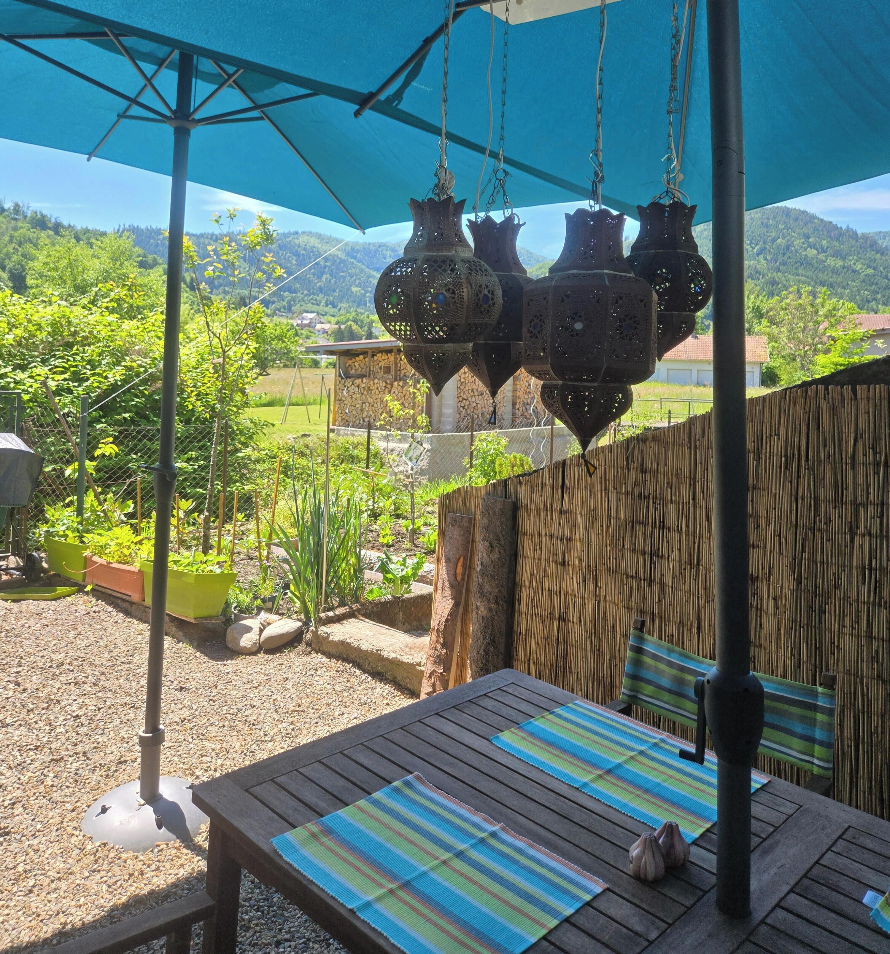 Een terras met een tafel en stoelen, een hanglamp, en uitzicht op een groen landschap met bomen en heuvels.