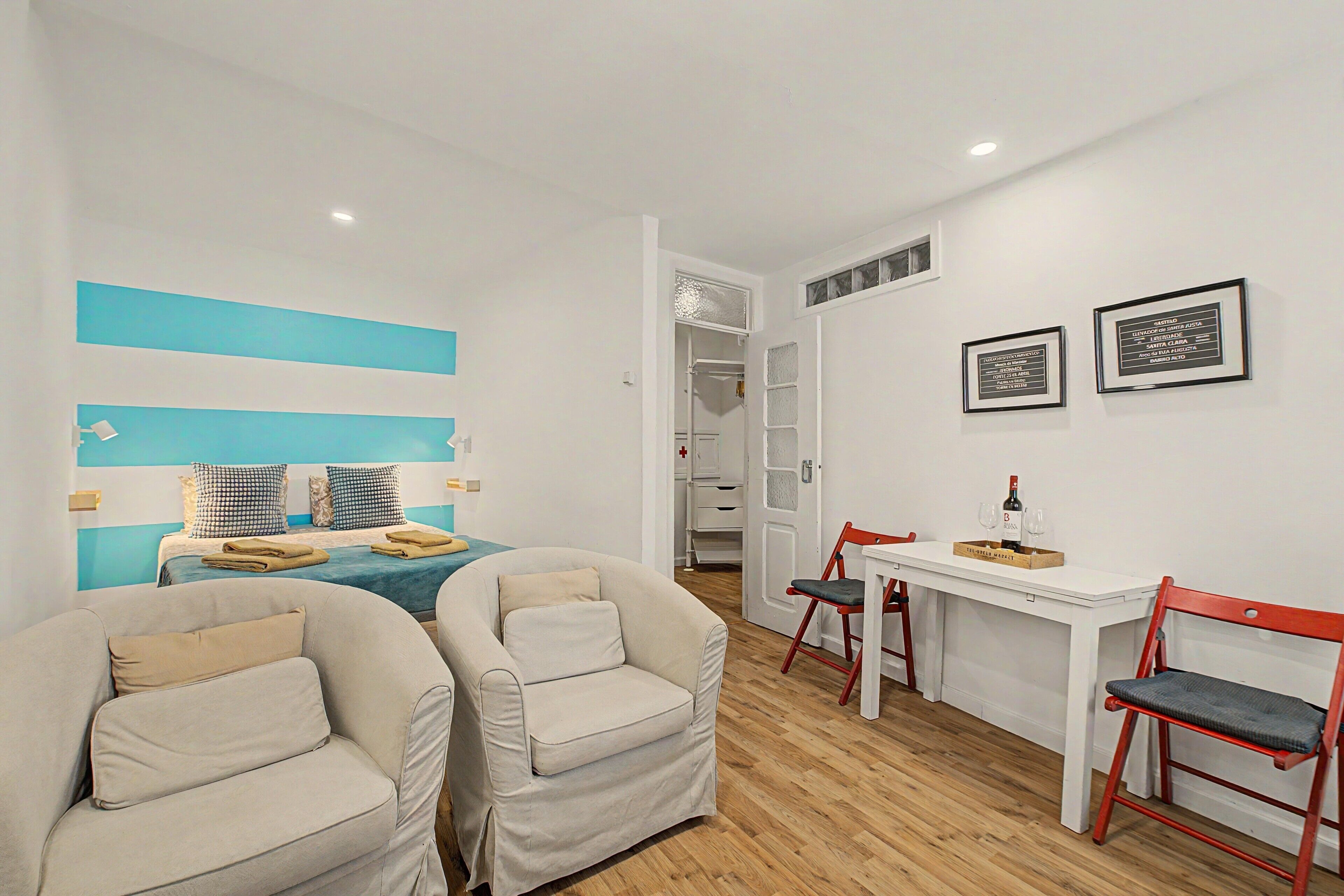 Une chambre d’hôtel avec un lit, deux fauteuils, une petite table avec une bouteille et des verres, et un mur orné de tableaux.