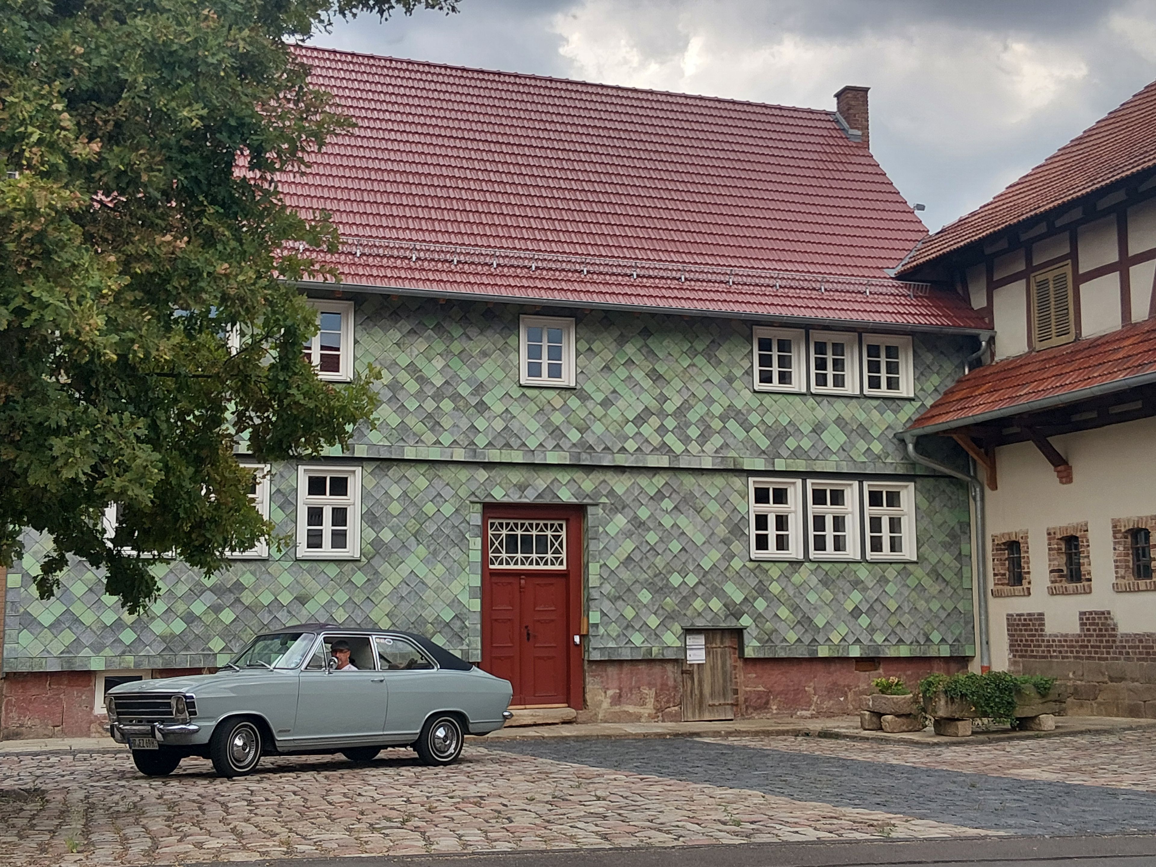 Ein zweistöckiges Gebäude mit rotem Ziegeldach, ein Auto vor der Tür und eine gepflasterte Straße.