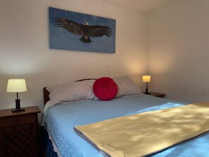 Un dormitorio con cama, dos lámparas de noche, una mesita de noche, un cojín rojo, una manta color beige y un cuadro de un águila en la pared.