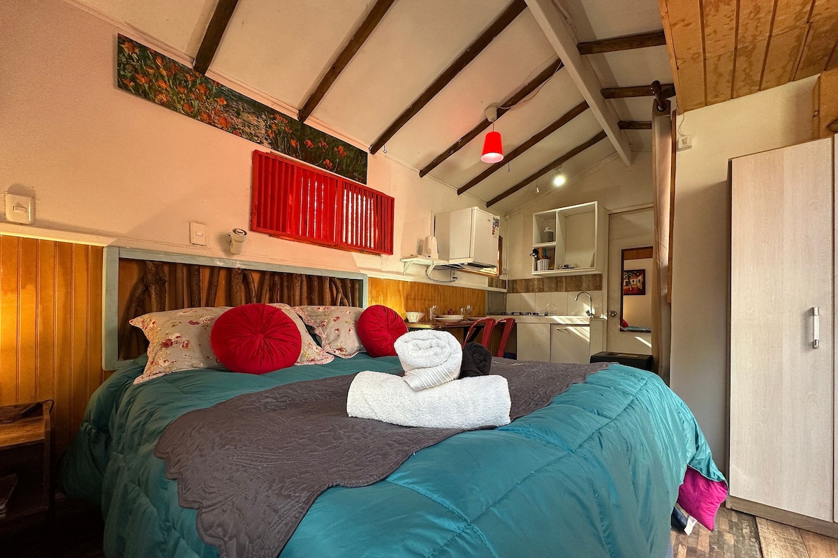 Un dormitorio con cama, almohadas, una colcha, cabecera roja, un cuadro, un espejo, un lavamanos y un armario.