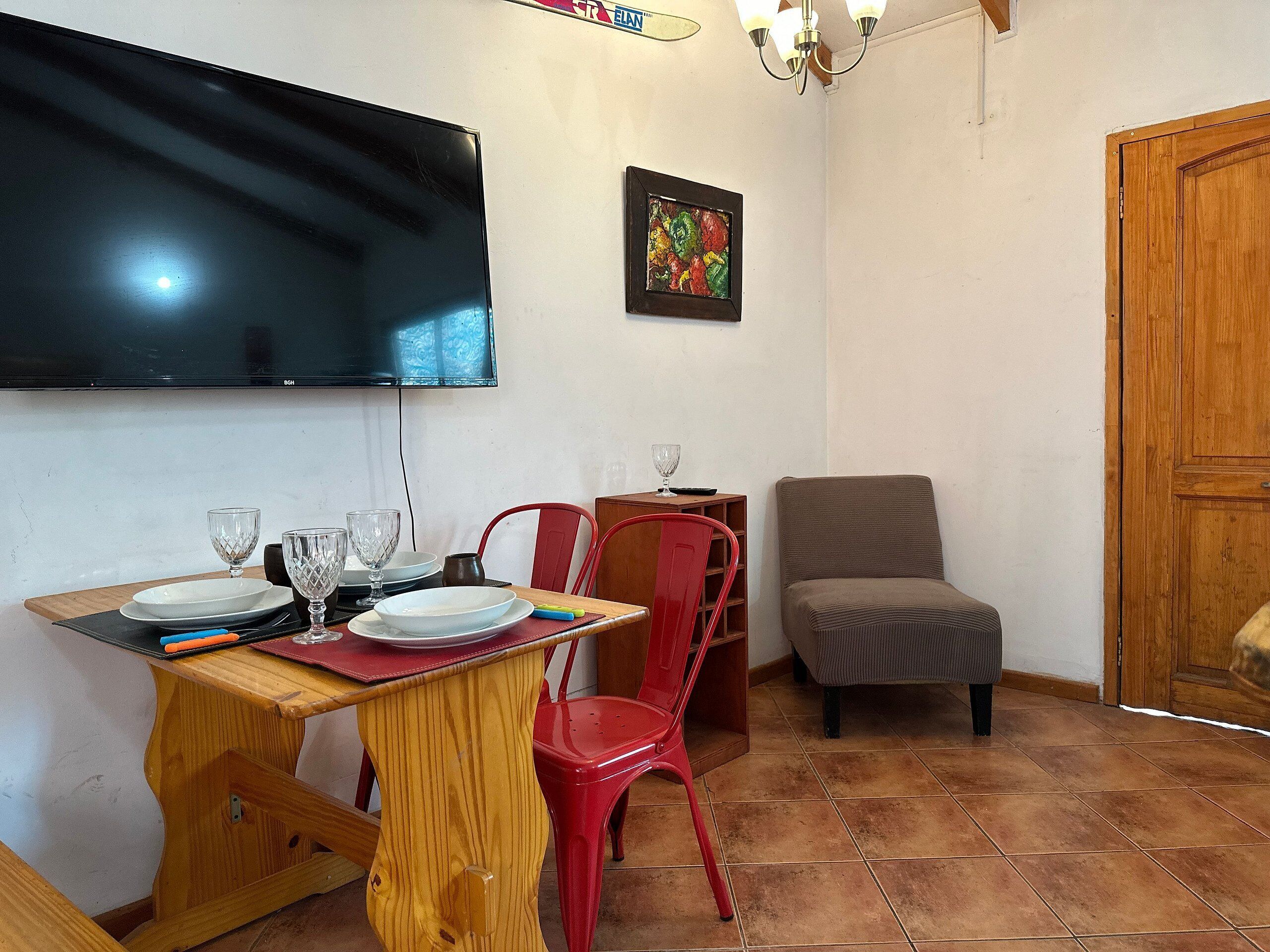 A dining area with a wooden table, red chairs, a television mounted on the wall, and a door to the right.