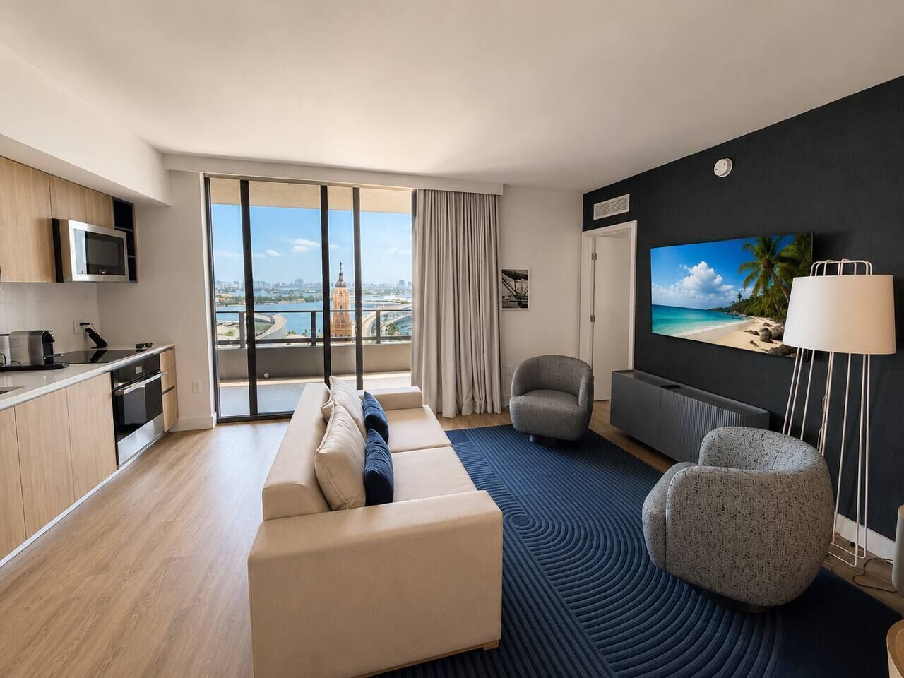A modern hotel room with a balcony, a sofa, a TV, a lamp, and a kitchen area.