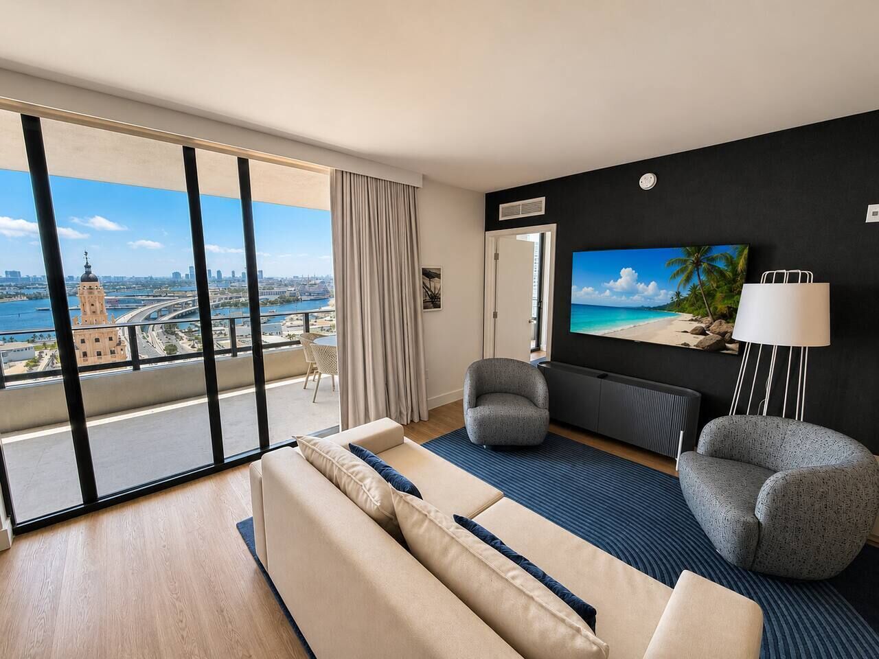 A modern hotel room with a large balcony, a sofa, a chair, a TV, and a lamp.