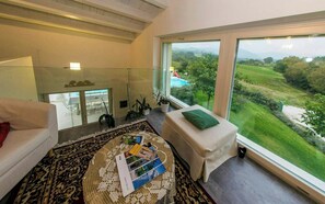 Un salon moderne doté d’une grande fenêtre donnant sur un paysage luxuriant, d’un canapé blanc, d’un petit pouf et d’un tapis à motifs.