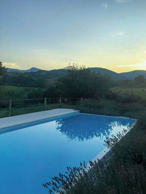 A rectangular swimming pool with a clear blue surface, surrounded by a wooden fence, set against a backdrop of rolling hills and a clear sky. - Wellness Retreat between Marche and Umbria (Sassoferrato)
