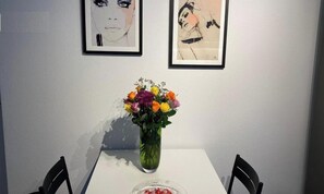 Ein Esstisch mit einer Blumenvase, zwei gerahmte Kunstwerke an der Wand und ein Stuhl an jeder Seite des Tisches.