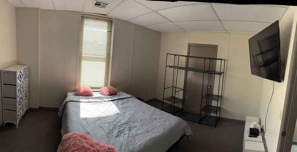 A bedroom with a bed, a wardrobe, a TV mounted on the wall, and a window with blinds.