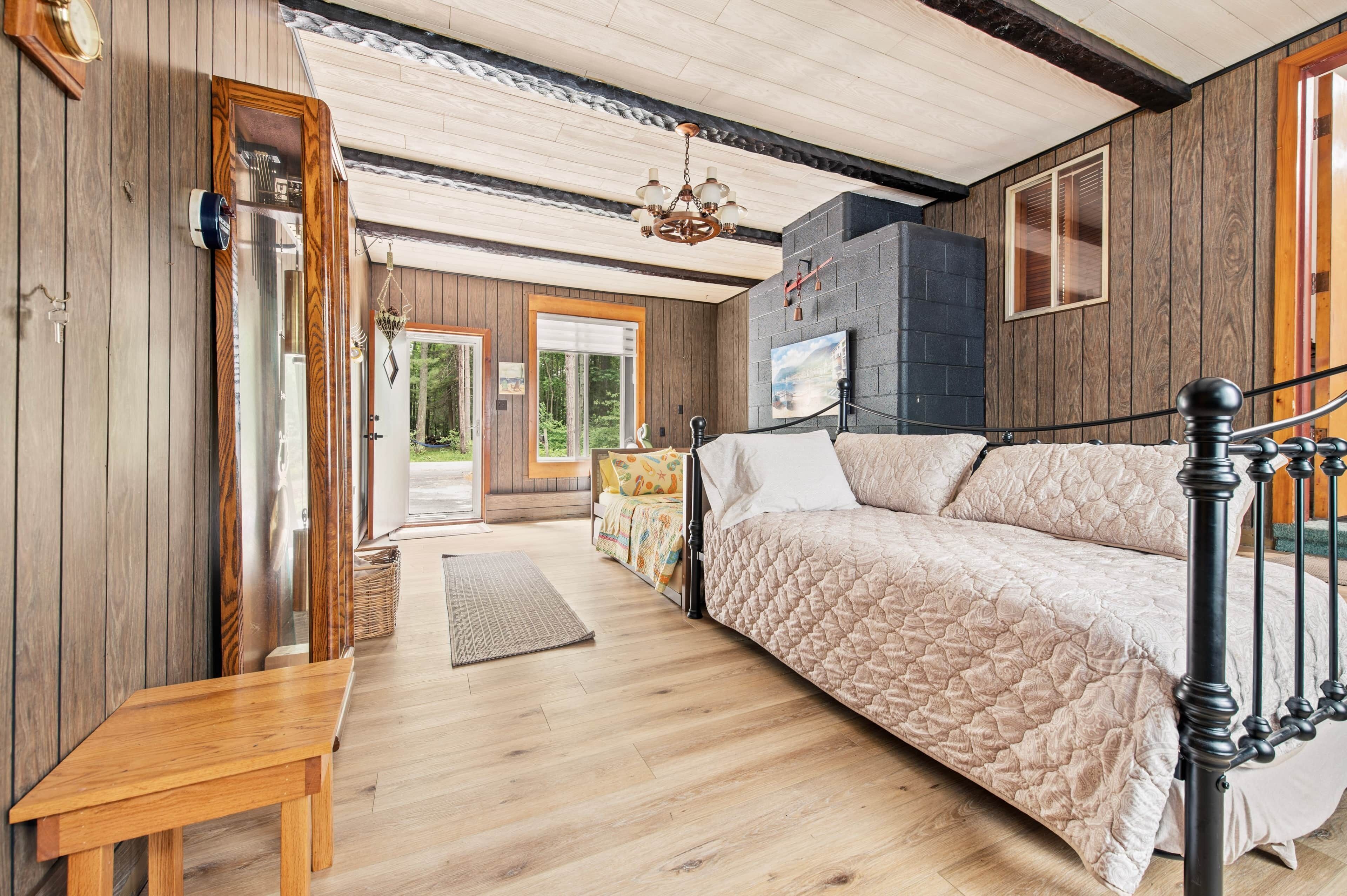 A bedroom with a wooden floor, a bed with a quilted cover, a wooden bench, a hanging light fixture, and a view of the outdoors through a door.