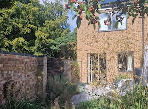 A brick building with a window and a door, surrounded by greenery and a brick wall.