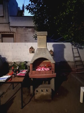 Ein Backofen aus Ziegelsteinen im Freien, in dem Essen zubereitet wird, ein Tisch mit Speisen und Getränken sowie eine an der Wand angelehnte Leiter.