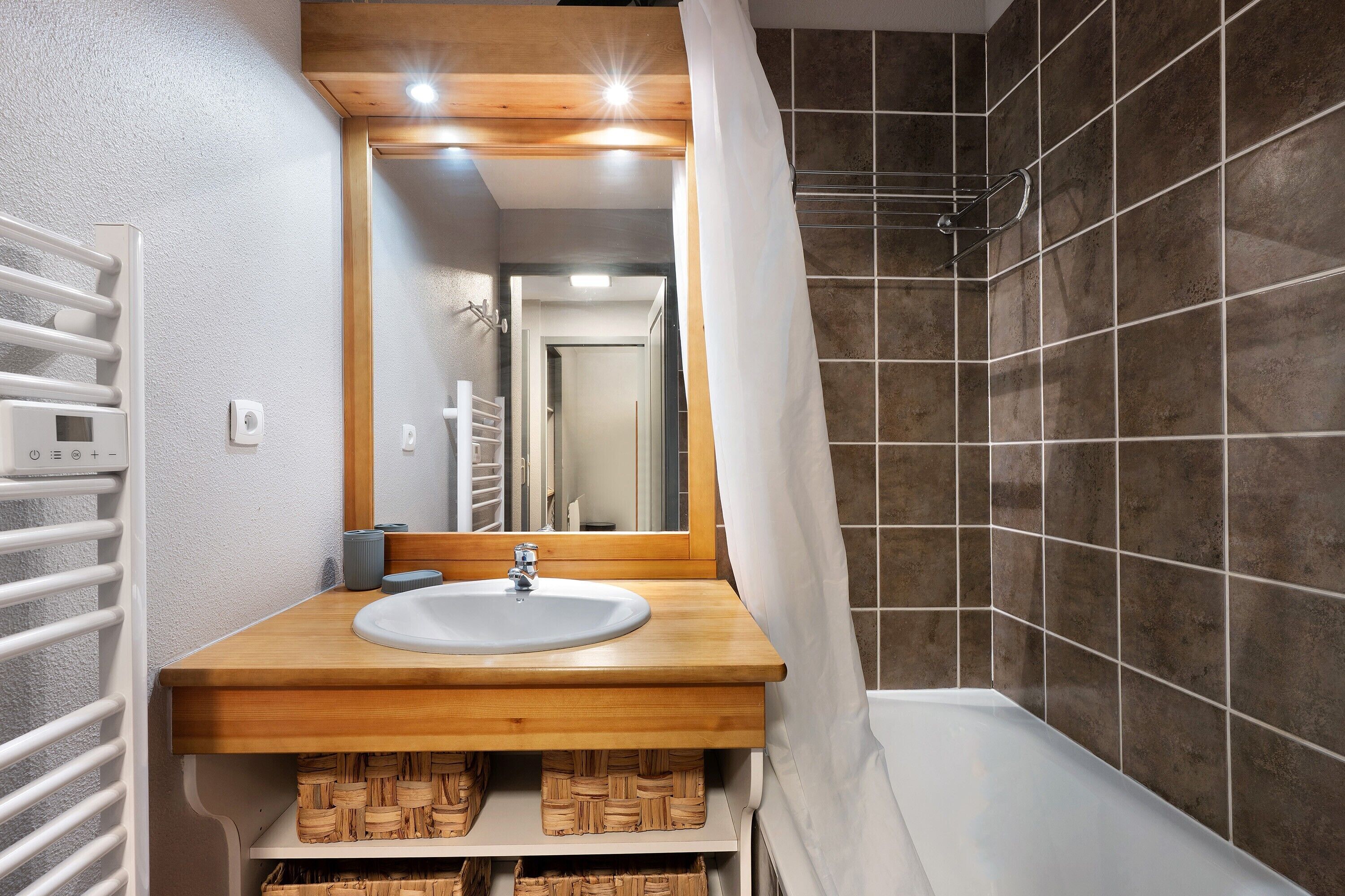 Une salle de bain moderne avec un meuble-lavabo en bois, un lavabo blanc et un espace douche aux carreaux foncés.