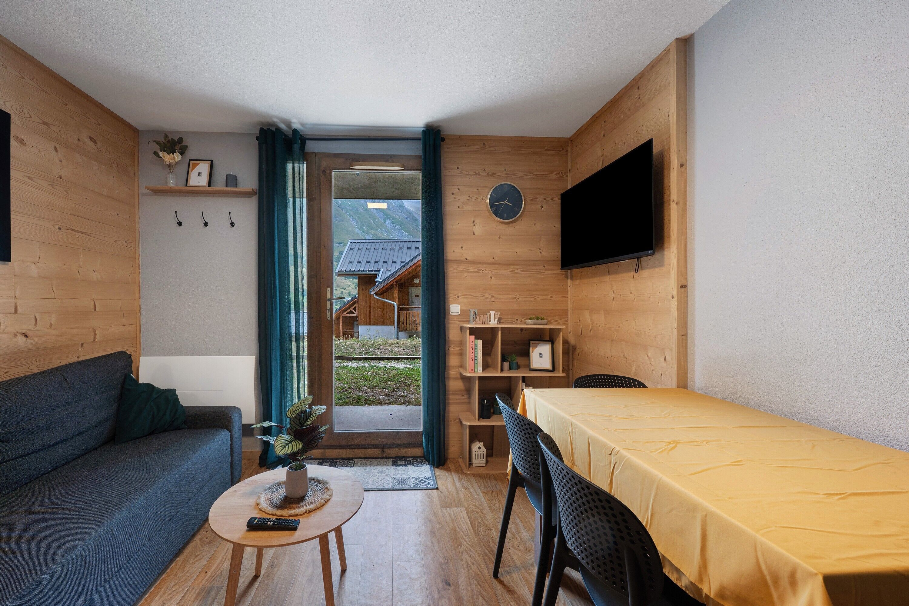 Une pièce chaleureuse avec un canapé, une petite table sur laquelle est posée une plante, un lit recouvert d’une couverture jaune, un téléviseur fixé au mur et une étagère en bois où sont rangés des livres et des objets décoratifs.