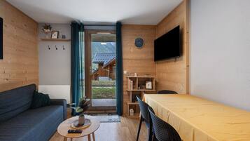 Une pièce chaleureuse avec un canapé, une petite table sur laquelle est posée une plante, un lit recouvert d’une couverture jaune, un téléviseur fixé au mur et une étagère en bois où sont rangés des livres et des objets décoratifs.
