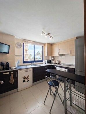 Een moderne keuken met een vaatwasser, een magnetron en een koelkast. Er is een raam met uitzicht op palmbomen en gebouwen.