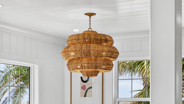 A room with white paneled walls, a large woven pendant chandelier, and large windows with views of palm trees. - Sandcastle Villa 3BR Beachfront Home (Clearwater Beach)