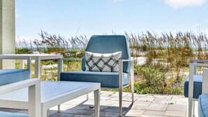 A patio with outdoor furniture, including a chair with a patterned cushion, a table, and a view of the beach with tall grasses. - Sandcastle Villa 3BR Beachfront Home (Clearwater Beach)