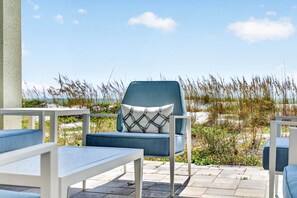 A patio with outdoor furniture, including a chair with a patterned cushion, a table, and a view of the beach with tall grasses. - Sandcastle Villa 3BR Beachfront Home (Clearwater Beach)