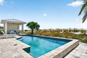 A rectangular swimming pool with a tiled deck, a covered seating area with chairs, and a palm tree. - Sandcastle Villa 3BR Beachfront Home (Clearwater Beach)