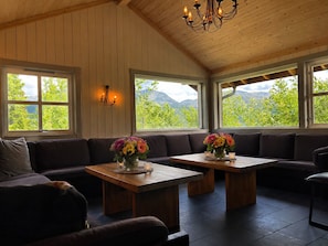Una habitación con techo de madera, ventanas grandes con vistas a la vegetación y montañas, un sofá y dos mesas con arreglos florales.