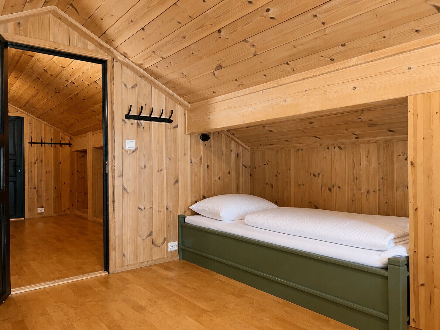 A wooden-paneled room with a single bed, a coat rack, and a door leading to another room.