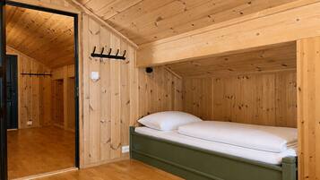 A wooden-paneled room with a single bed, a coat rack, and a door leading to another room.