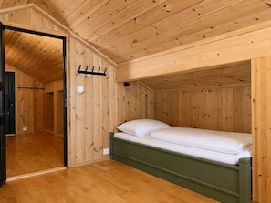 Una habitación con paneles de madera, una cama individual, un perchero y una puerta que da a otra habitación.