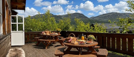 An outdoor wooden deck with picnic tables and benches, surrounded by greenery and mountains in the background.