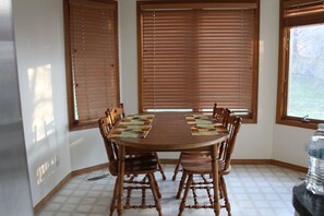 Uma área de refeições com mesa e cadeiras de madeira, janelas amplas com persianas e piso de azulejos.