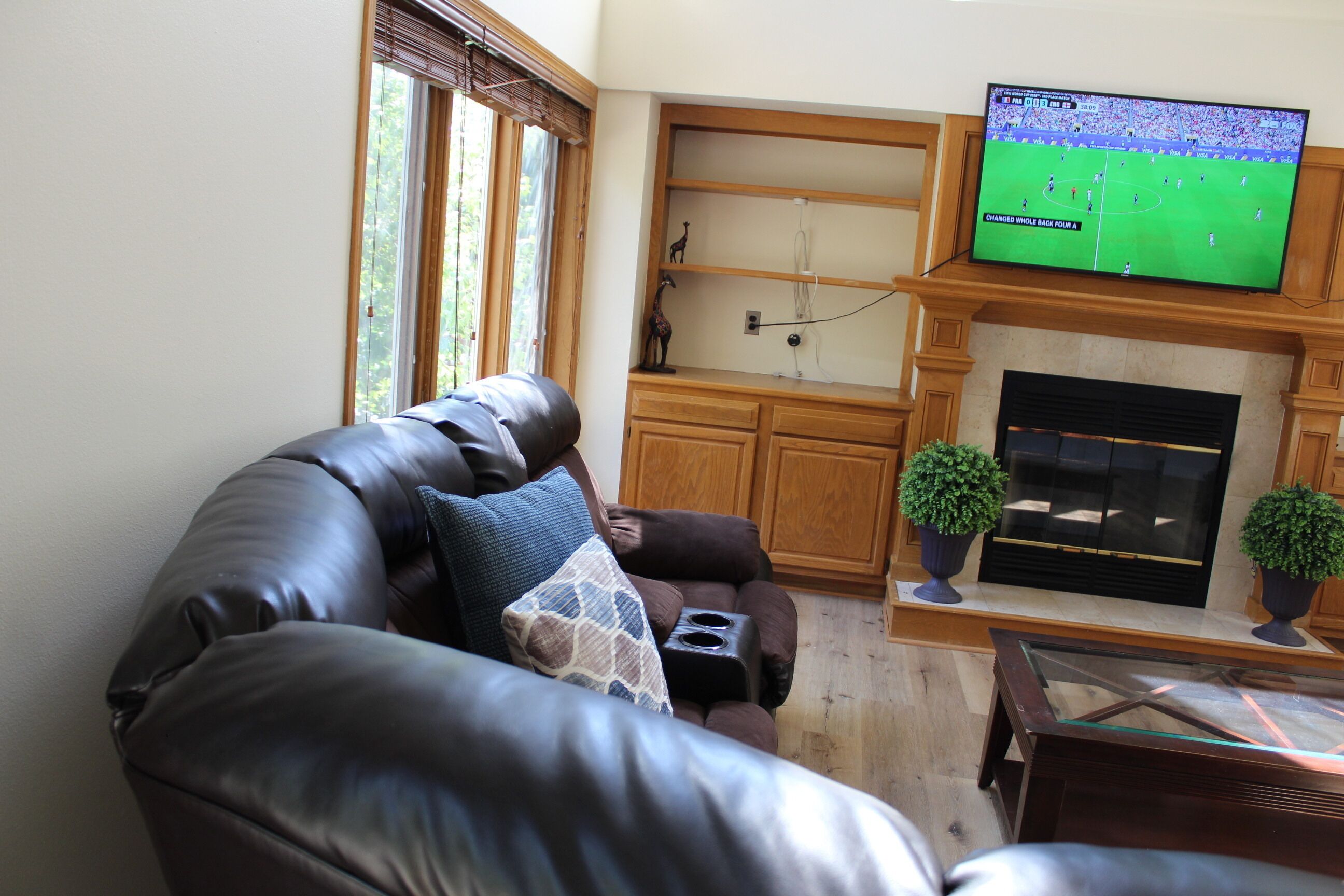 A living room with a leather sofa, a flat-screen TV mounted above a fireplace, and a window with blinds.
