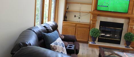 A living room with a leather sofa, a flat-screen TV mounted above a fireplace, and a window with blinds.