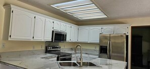 A kitchen with white cabinets, a stainless steel refrigerator, a microwave, and a sink. - The Johnston Paradise (Johnston)