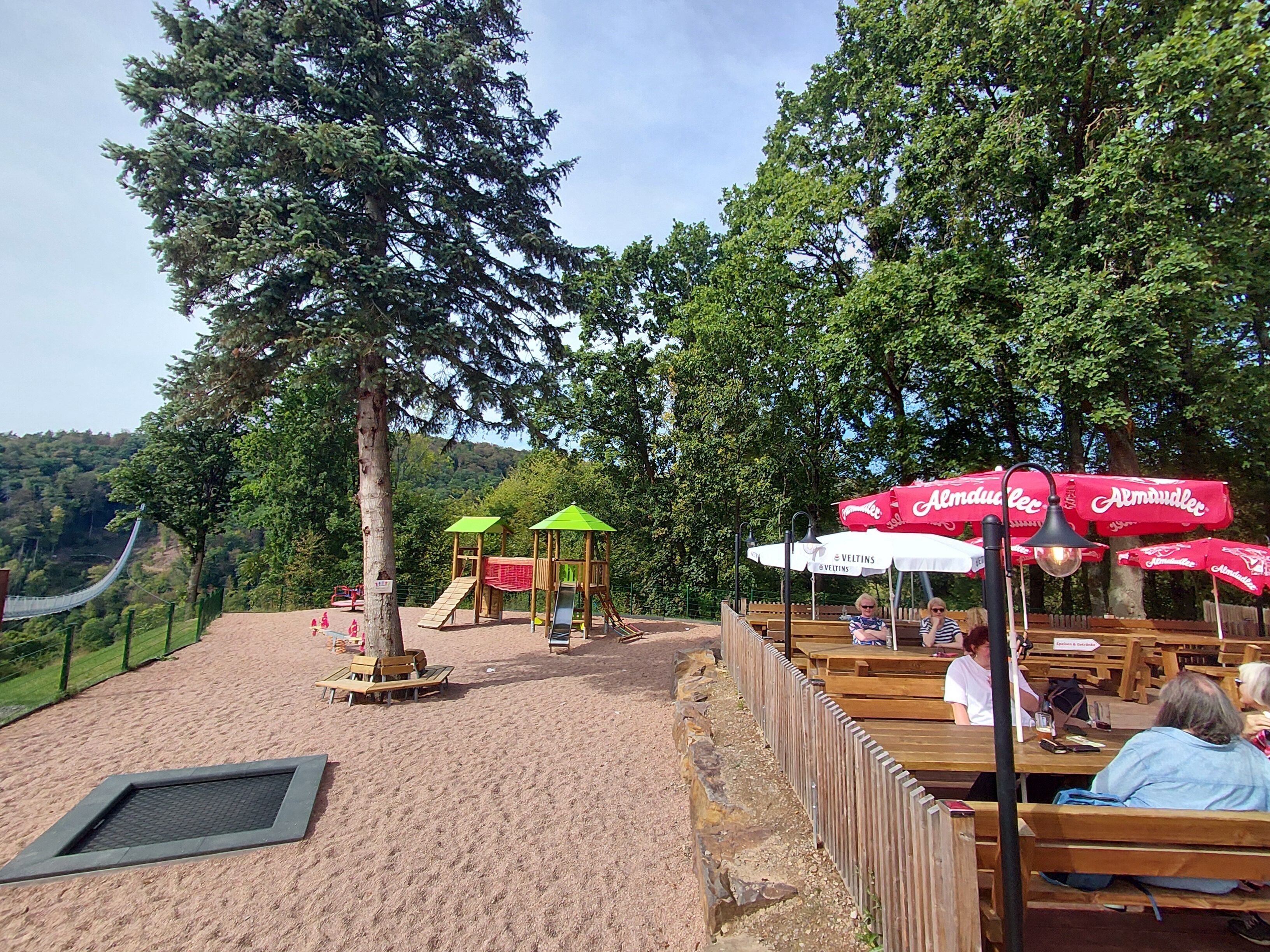 Ein Park mit Holzbänken, einem Spielplatz und roten Sonnenschirmen.