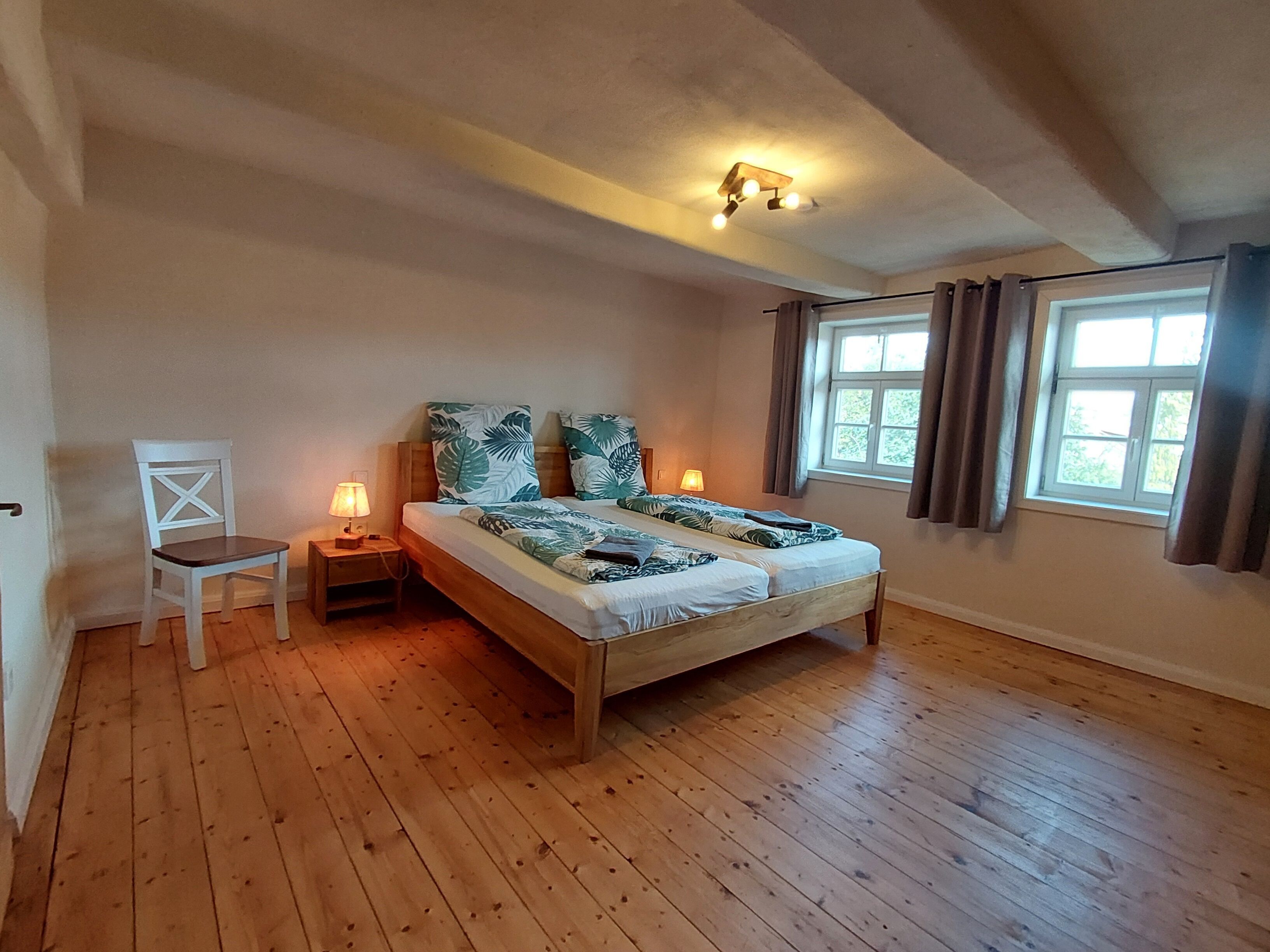Quarto com cama de madeira, duas luminárias de cabeceira, uma cadeira e janela com cortinas.